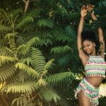 A joyful young woman posing in colorful attire amidst lush greenery outdoors.